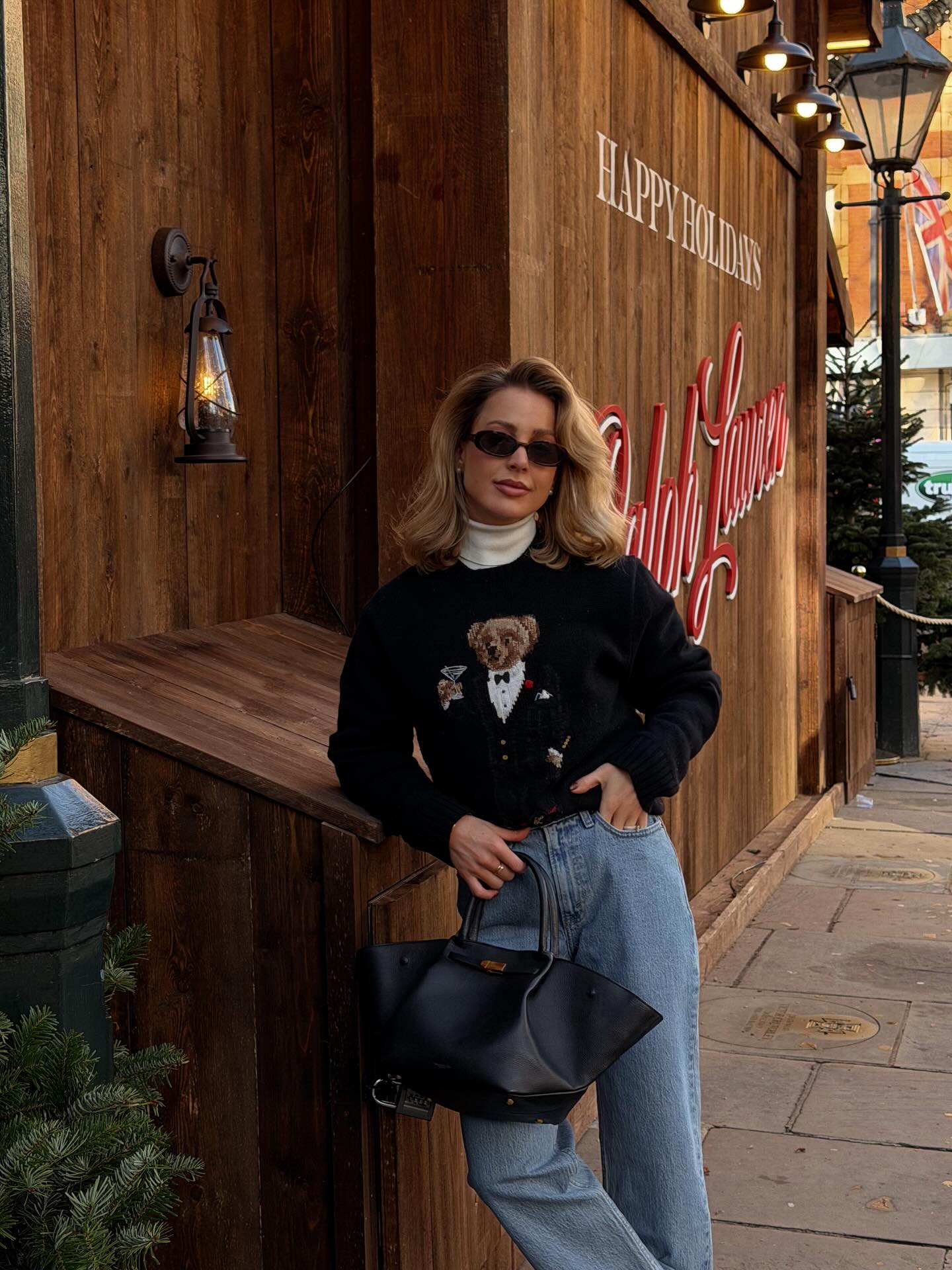 Casual holiday sweater outfit with a black teddy bear sweater layered over a white turtleneck, light-wash jeans, and a black tote bag.