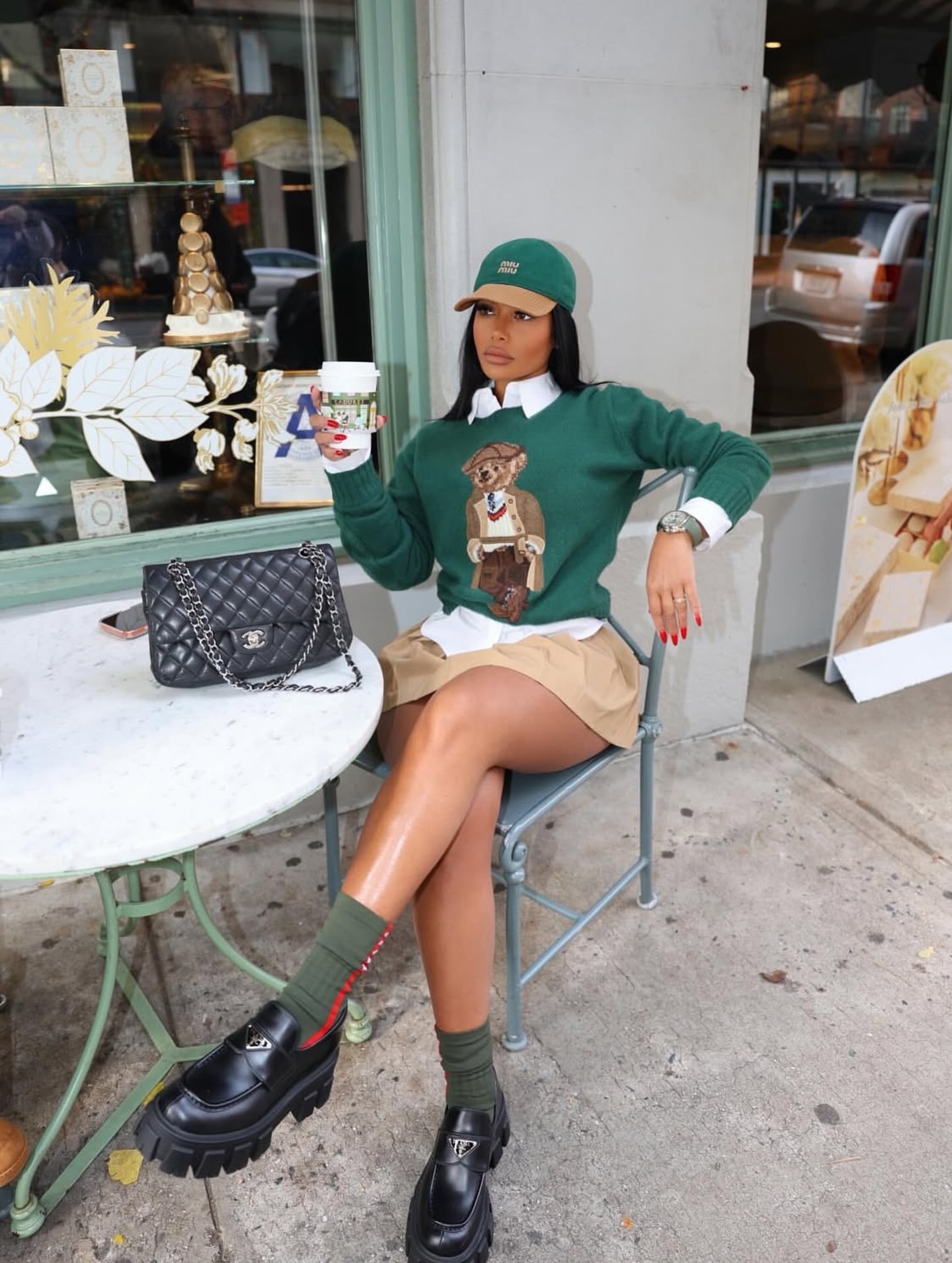 Preppy holiday sweater outfit with a green teddy bear knit, white collared shirt, khaki mini skirt, green cap, and chunky black loafers.