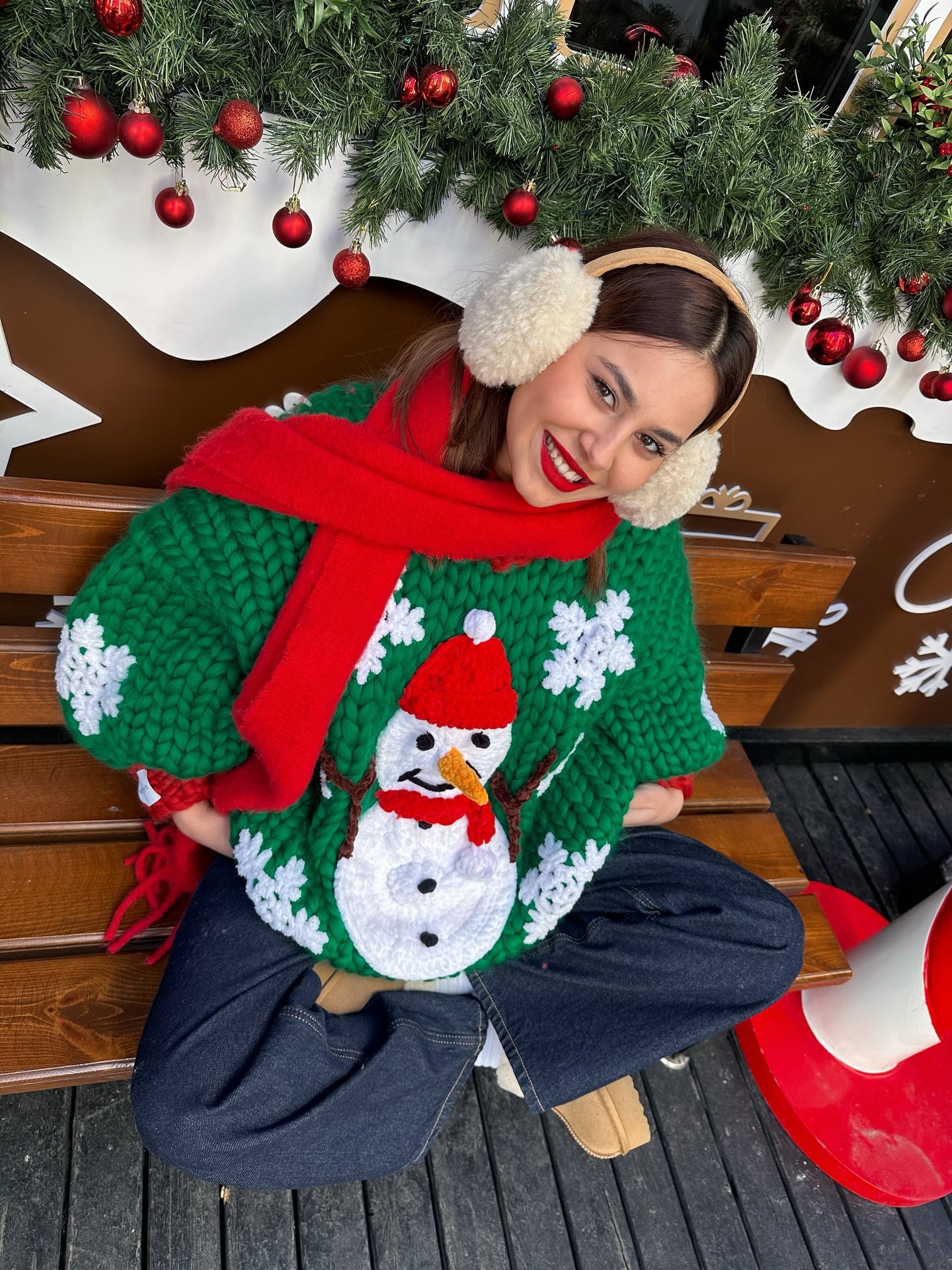Ugly Christmas sweater outfit with a chunky green snowman sweater, relaxed dark jeans, red scarf, fluffy earmuffs, and tan UGG boots.