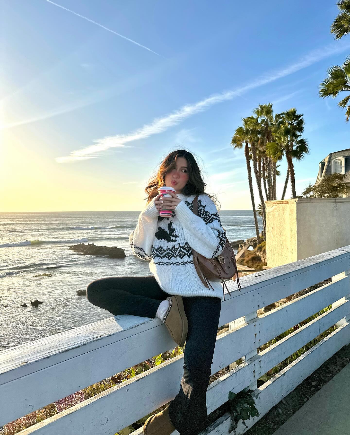 Casual Christmas sweater outfit with a white and black reindeer knit, black pants, tan UGG boots, and a brown shoulder bag.