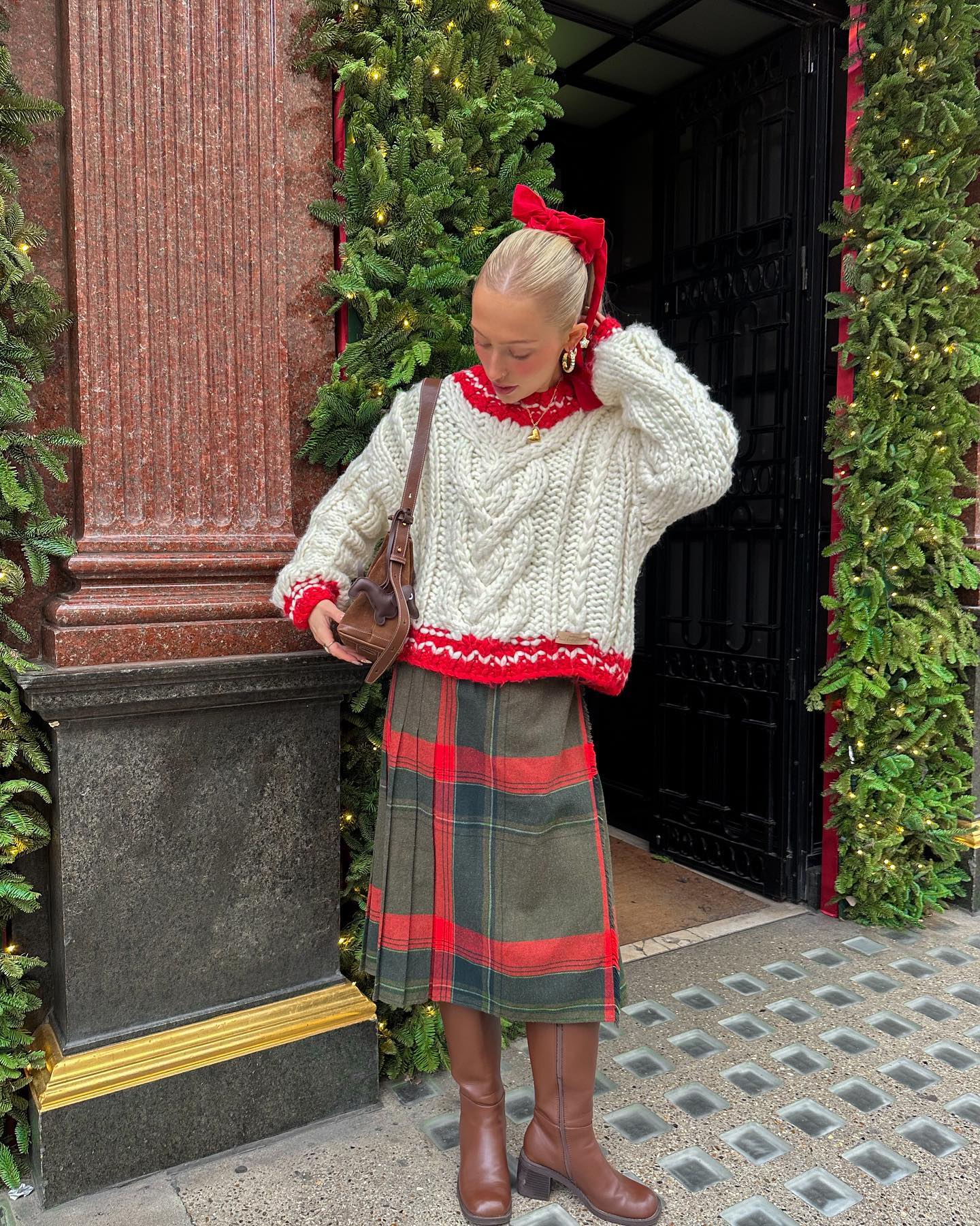Christmas sweater outfit with a cream cable knit sweater with red trim, red and green plaid midi skirt, and brown leather boots.
